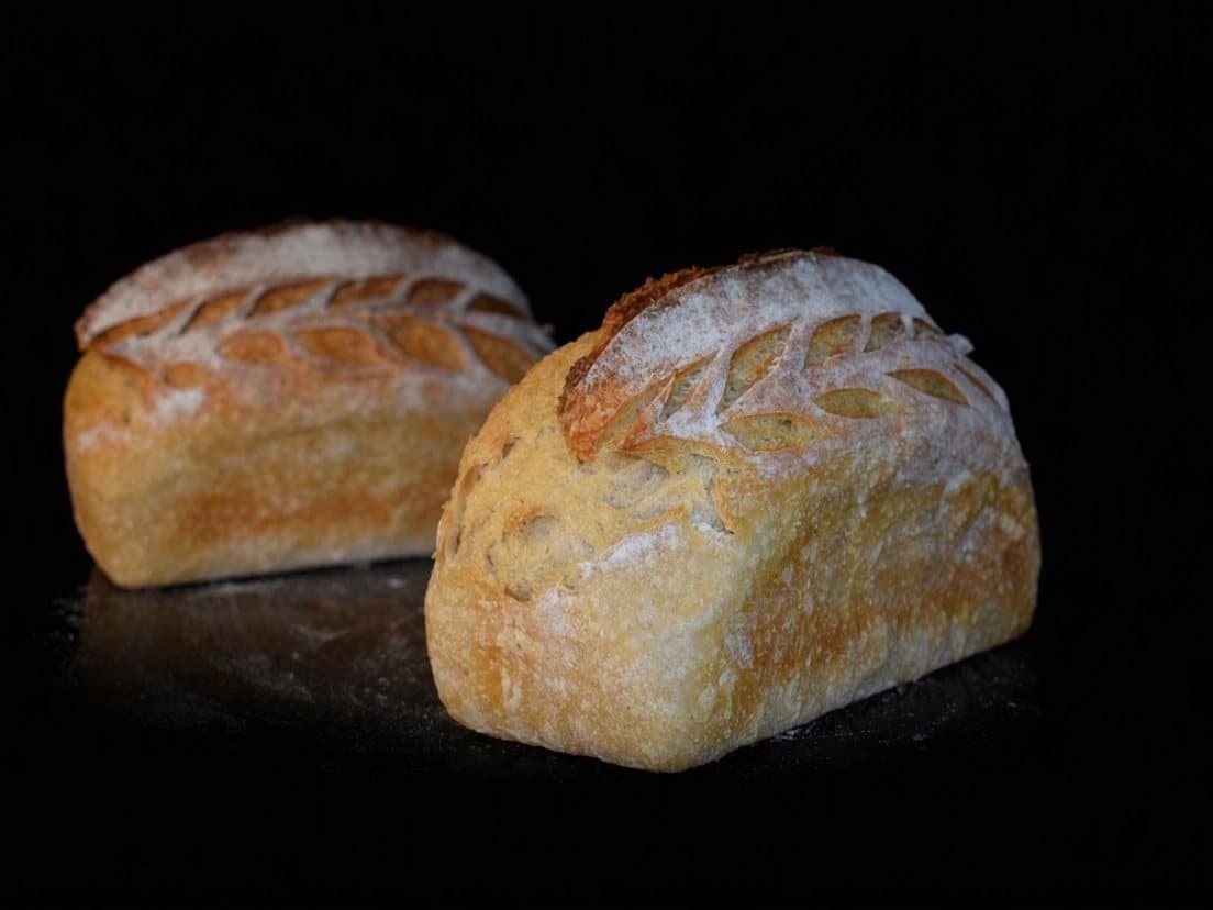 Two white sourdough loaves with a scored ear of wheat and flour on the crust, on a black counter.
