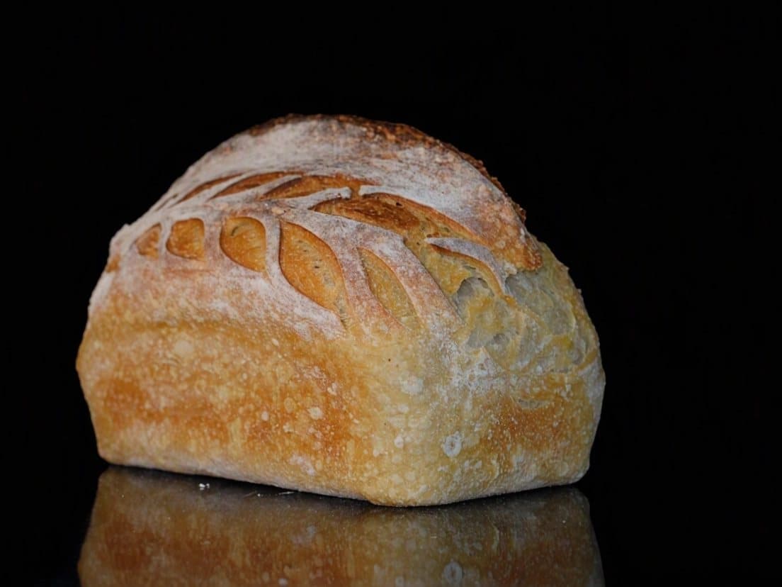 A white sourdough loaf with a golden crust and an ear of wheat scored into it, on a black counter.