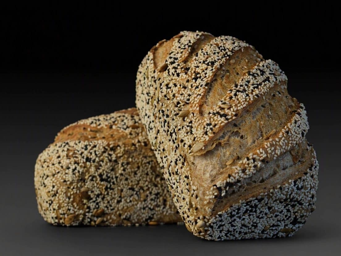 Two dark loaves covered in white and black sesame seeds, against a dark background.